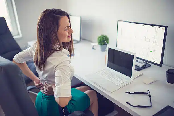 Young woman having back pain while sitting at desk in office.