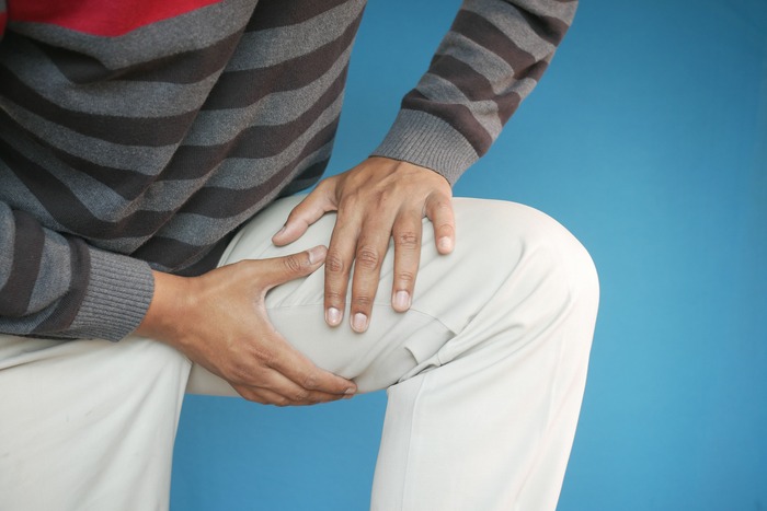 Close-up of a man's hands on his thigh, showing pain or injury, on a blue background